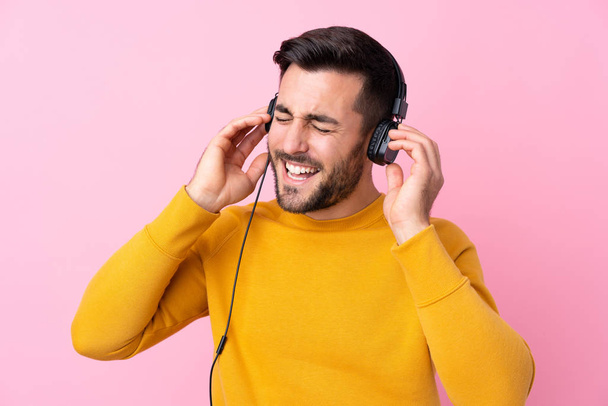 Jonge knappe man met baard luisterend naar muziek over geïsoleerd roze - Foto, afbeelding