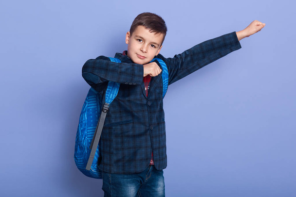 Image of confident joyful young boy raising arms, making gesture, looking directly at camera, getting pleasure from study, standing isolated over lilac background in studio. Studying concept. - Foto, Imagem