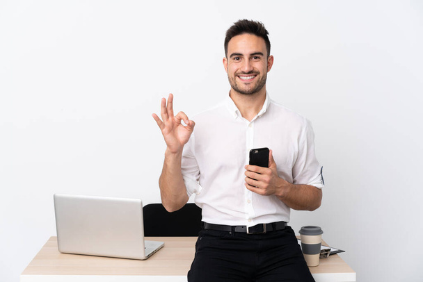 Молодой бизнесмен с мобильным телефоном на рабочем месте показывает знак ОК с пальцами
 - Фото, изображение