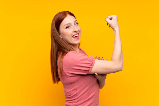 Mladý ryšavý žena přes izolované žluté pozadí dělat silné gesto - Fotografie, Obrázek