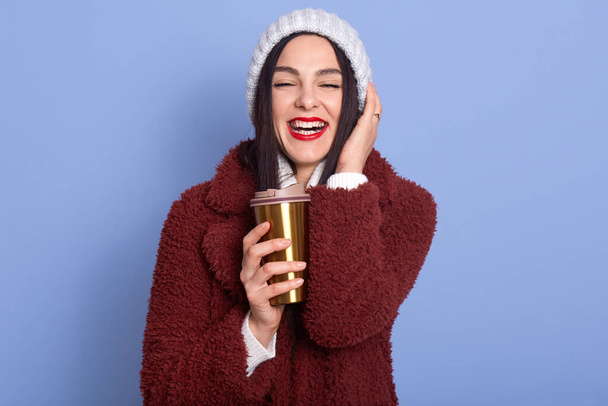 Horizontal shot of darkhaired girl with winter hat posing isoalted over blue background, holds coffee in thermo mug, moves towards zero waste, female looks happy, looking directly at camera with smile - Fotografie, Obrázek