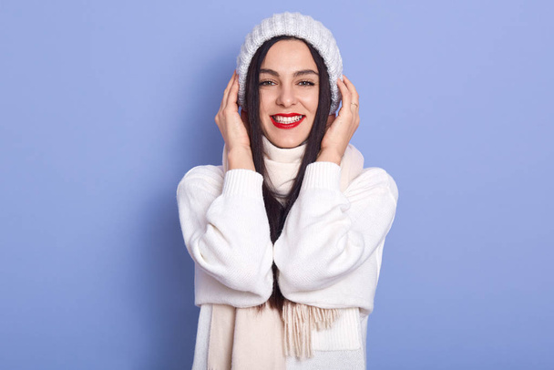 Image of cheerful magnetic good looking young female looking directly at camera, wearing white sweater, scarf and hat, putting hands on head, having red lips, smiling sincerely. Emotions concept. - Photo, Image