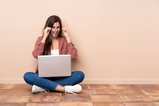 Adolescente estudiante chica sentada en el suelo con un ordenador portátil con gafas y sorprendida
 - Foto, Imagen