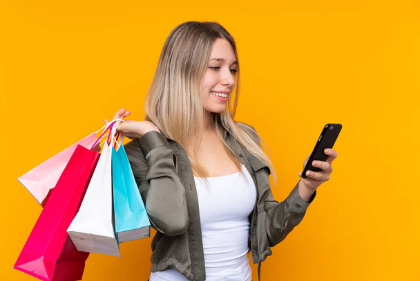 Young blonde woman over isolated yellow background holding shopping bags and writing a message with her cell phone to a friend - Foto, imagen