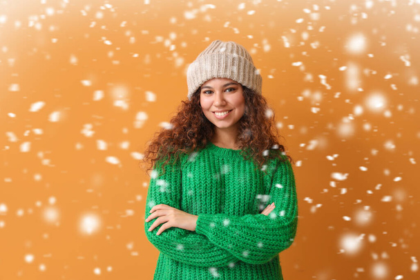 Young African-American woman in warm sweater and falling snow on color background - Foto, Imagem