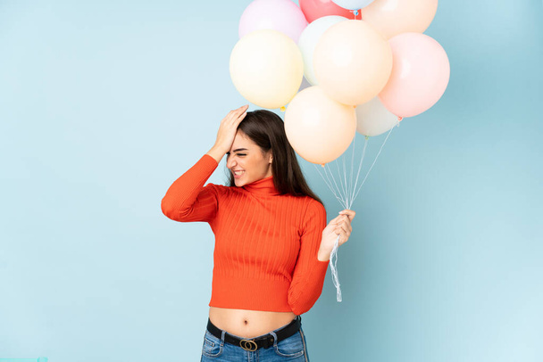 Junge Frau fängt viele Luftballons isoliert auf blauem Hintergrund und hat Zweifel mit verwirrtem Gesichtsausdruck - Foto, Bild