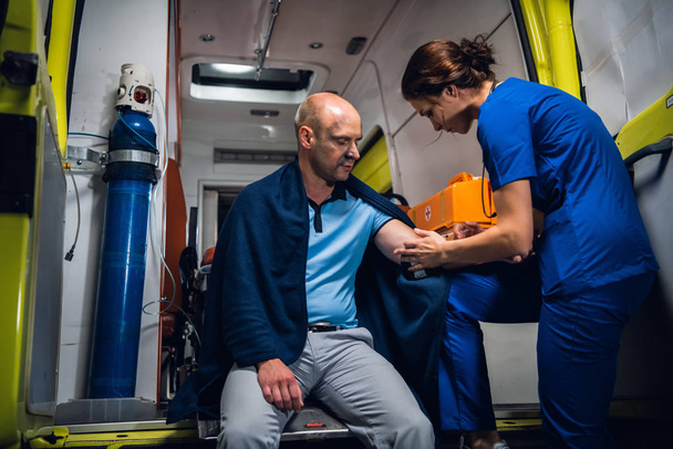 El hombre está sentado en manta, la enfermera de uniforme hace una inyección en el coche de la ambulancia
 - Foto, Imagen