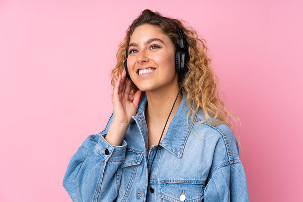 Young blonde woman with curly hair isolated on pink background listening music - Valokuva, kuva