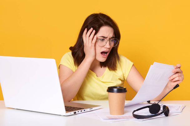 Studio shot of astonished woman looking at documents in her hand, sitting at her work desk, wearing casually, working at home, isolated on yellow background, holds paperswith widely opened mouth. - Photo, Image