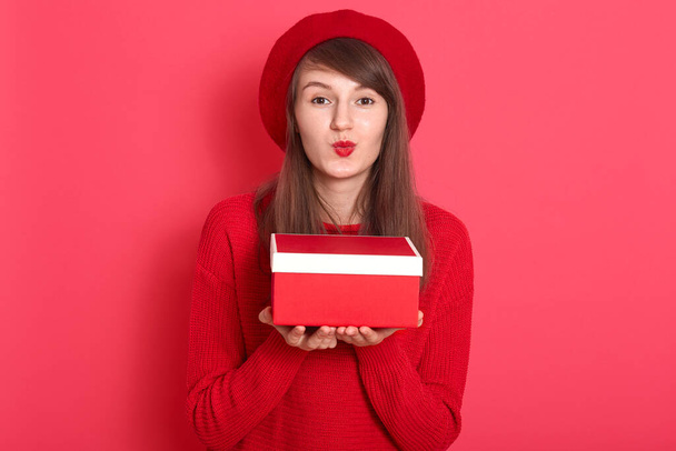 Close up portrait of beautiful woman wearing red sweaterand beret, holding in hand red gift box, making kiss gesture, wants to give present for her boyfriend, posing isolated over studio wall. - Foto, immagini