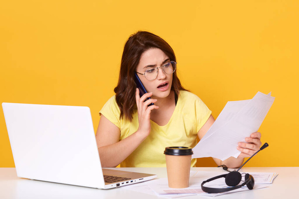 Close up portrait of business woman working online via laptop, isolated over yellow studio background, talking to somebody via smart phone, holding paper in hands, looks astonished. and concentrated. - Foto, afbeelding