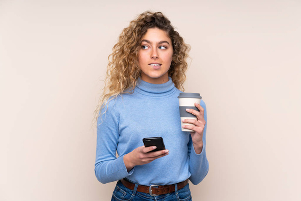 Nuori blondi nainen kihara tukka yllään poolopaita eristetty beige tausta tilalla kahvia ottaa pois ja mobiili kun ajattelee jotain
 - Valokuva, kuva