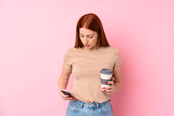 Fiatal vörös hajú nő izolált rózsaszín háttér kezében kávé elvitelre, és egy mobil - Fotó, kép