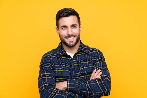 Young handsome man with beard over isolated yellow background keeping the arms crossed in frontal position - Zdjęcie, obraz