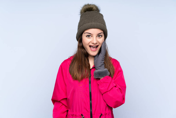 Молода дівчина з зимовим капелюхом ізольована на синьому фоні з несподіваним і шокованим виразом обличчя
 - Фото, зображення
