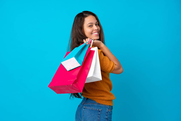 Young Brazilian girl over isolated blue background holding shopping bags and smiling - Foto, imagen