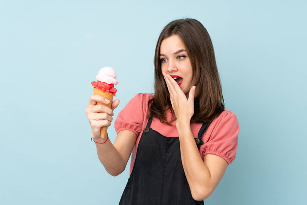Дівчина-підліток тримає корнет морозиво ізольоване на синьому фоні з несподіваним і шокованим виразом обличчя
 - Фото, зображення