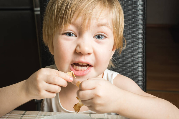 Beautiful baby with blue eyes eats delicious pancakes while sitting at kitchen table on chair. - Photo, image