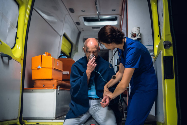 Man zit in deken met zuurstofmasker, dokter in medisch uniform houdt zijn hand vast - Foto, afbeelding