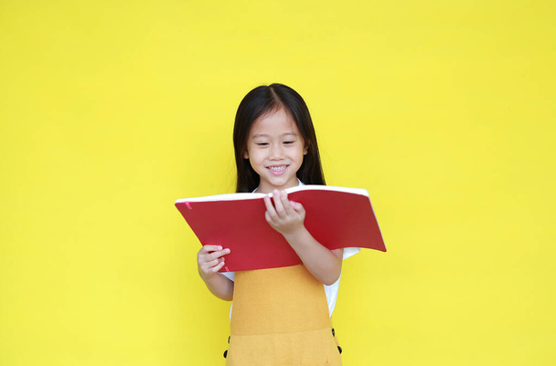 Asian little child girl reading book isolated on yellow background. Portrait of schoolgirl smiling while study. Back to School and education concept - Photo, Image