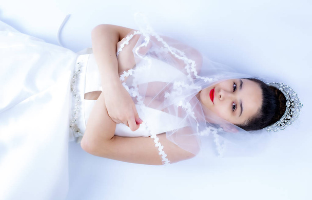 Beautiful young bride in white wedding dress and veil indoors. Luxuty model lying over white backgroung. Girl waiting her groom in bright room. Beautiful wedding hairdress. High key phot - Fotografie, Obrázek
