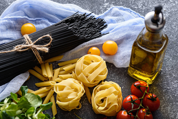 ingredients for dinner-black spaghetti on pastel lilac napkin , fettuccine, tomatoes, corn salad, olive oil on gray background - Photo, Image