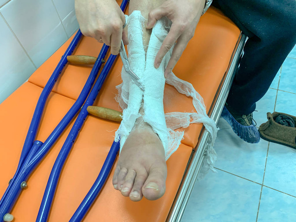 Молодой человек снимает гипс с ноги ножницами в больнице. Костыли лежат неподалеку
 - Фото, изображение