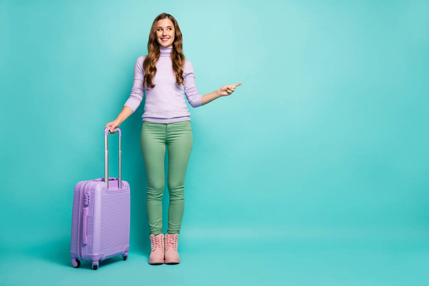 Full body photo of funny lady airport with rolling trip case direct finger empty space choose best airlines wear lilac sweater green trousers footwear isolated pastel teal color background - Fotoğraf, Görsel