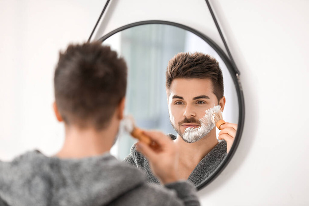 Όμορφος νεαρός άνδρας ξυρίζεται κοντά στον καθρέφτη στο μπάνιο - Φωτογραφία, εικόνα