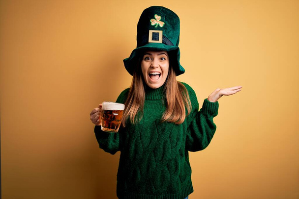 Joven hermosa mujer con sombrero verde bebiendo tarro de cerveza en la celebración del día de San Patricio muy feliz y emocionada, expresión ganadora celebrando la victoria gritando con una gran sonrisa y las manos levantadas
 - Foto, imagen