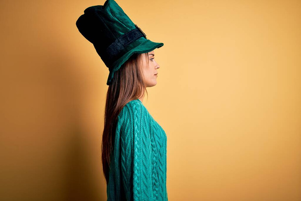 Giovane bella donna bruna con cappello verde con trifoglio che celebra giorno santo patricks cercando di lato, rilassarsi posa profilo con il viso naturale con sorriso fiducioso
. - Foto, immagini