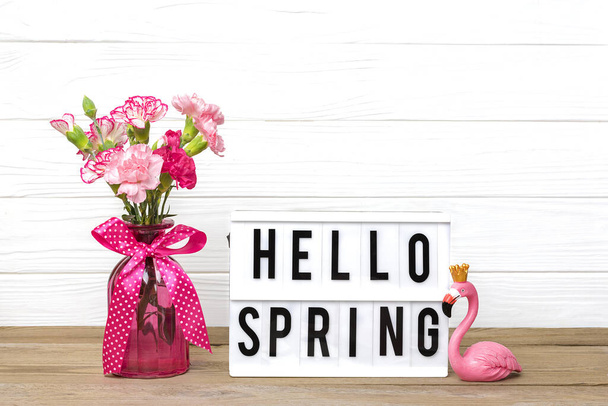 Маленькие цветные розовые гвоздики в вазе и лайтбоксе с текстом Hello Spring, фигурка фламинго на белом деревянном фоне Holiday Card
 - Фото, изображение