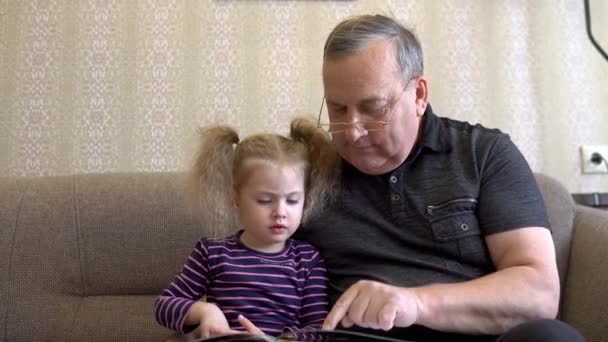 Enkelin liest mit Großvater ein Buch. Das Mädchen runzelt die Stirn und hört Großvater aufmerksam zu. Gemeinsam auf der Couch sitzen. - Filmmaterial, Video