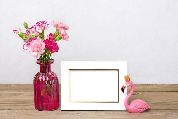 Small colored pink carnations in a vase and white photo frame, a figure of flamingo on wooden background and grey wall Hello spring, seasonal concept Mock up Place for text - Photo, Image