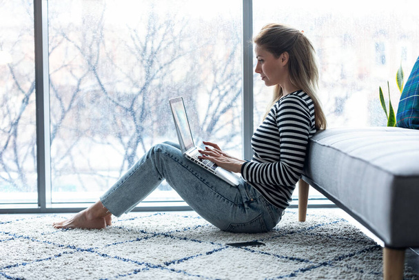 Pretty young woman using her laptop while sitting on the floor at home. - Фото, изображение