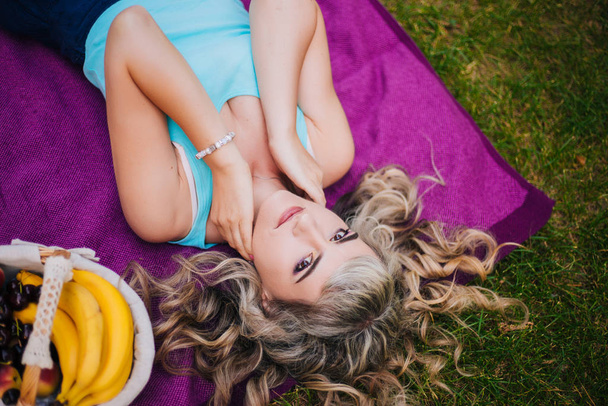 Gyönyörű lány piknikezik a parkban. Szőke, lila ágytakarón pihen. Modell, vidám mosollyal - Fotó, kép