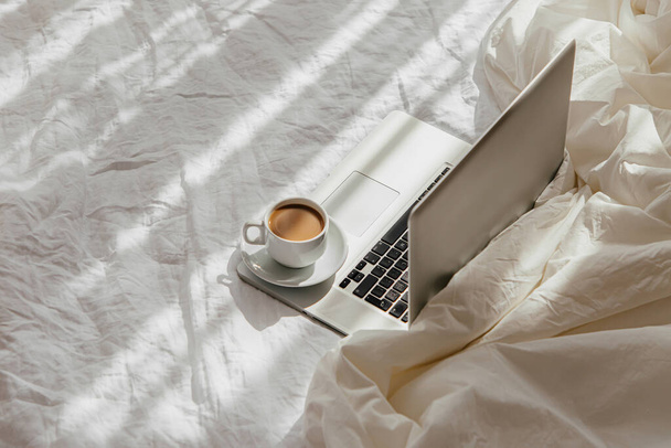 Laptop and cup of coffee on white bed with a  blanket. Work at home concept. Morning light  - Photo, image