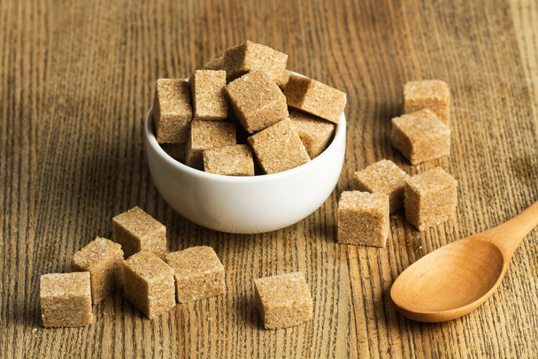 Кубики коричневого цукру в білій мисці і на дерев'яному столі
 - Фото, зображення