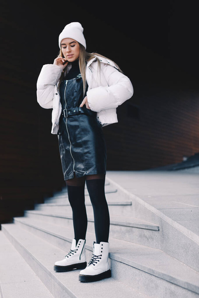 Rubia de pelo largo, con una gorra de lana blanca, una chaqueta blanca con un vestido curvilíneo de cuero negro debajo, medias negras y botas blancas, caminando elegantemente por un tramo de escaleras
 - Foto, Imagen