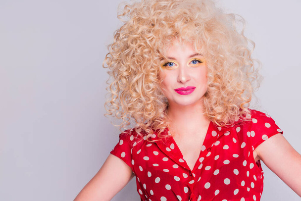 schönes blondes Mädchen mit lockiger Frisur, leuchtend gelbem Make-up und roter Tupfenbluse blickt in die Kamera.  - Foto, Bild