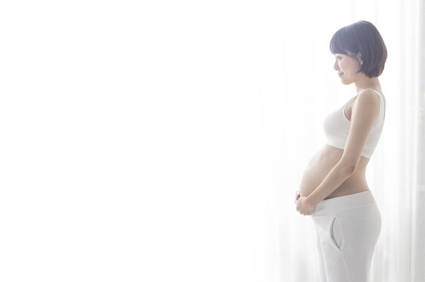 Молодая беременная азиатка стоит на полоборота и держится за живот.. - Фото, изображение