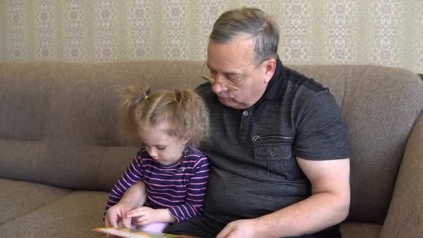 La nieta está leyendo un libro con el abuelo. La chica frunce el ceño ante el libro y escucha con atención al abuelo. Sentados en el sofá juntos de cerca - Imágenes, Vídeo