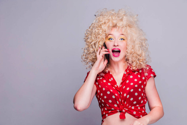 Beautiful blonde girl with a lush curly hairstyle, bright yellow makeup in her eyes and in a red polka-dot blouse is talking on the phone and is pleasantly surprised - Φωτογραφία, εικόνα