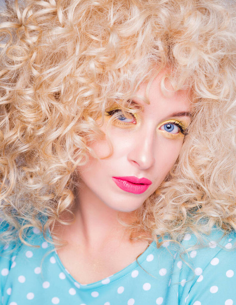 schöne modische blonde Mädchen im Retro-Stil mit voluminöser Lockenfrisur, in einer blauen Tupfenbluse auf grauem Hintergrund blickt in die Kamera.  - Foto, Bild