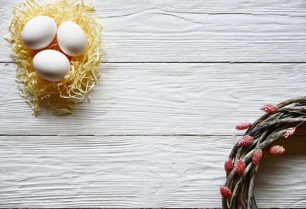 Гніздо з білими курячими яйцями і ротанговим вінком з сухими квітами на білому дерев'яному тлі. Вид зверху. Яскрава весна Великдень фон
. - Фото, зображення