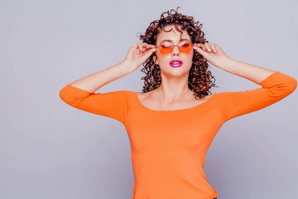Chica morena de moda en un suéter naranja brillante y gafas posando sobre un fondo gris
 - Foto, Imagen