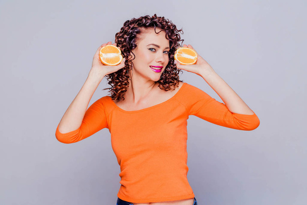 Όμορφη μελαχρινή κοπέλα με μακιγιάζ και σγουρό χτένισμα σε πορτοκαλί πουλόβερ ποζάροντας σε γκρι φόντο με δύο μισά του πορτοκαλιού - Φωτογραφία, εικόνα