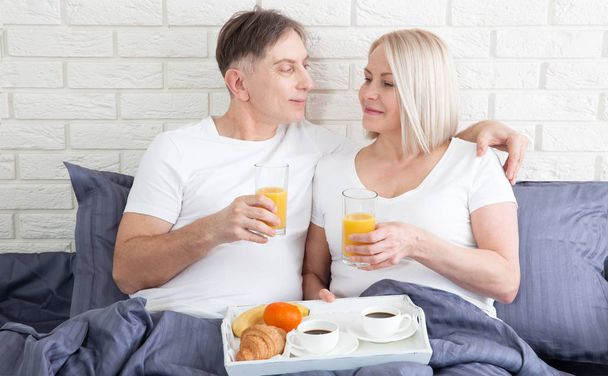 Beautiful couple having breakfast lying in bed at home. Handsome man and attractive woman are enjoying spending time together while gently hugging and kissing sitting in bed. - Foto, imagen