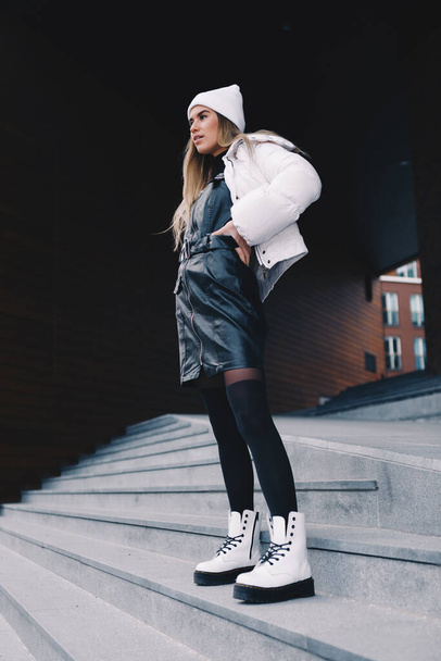 Long haired blonde, wearing a white woollen cap, a white jacket with a curvaceous black leather dress underneath, black stockings and white boots, walking gracefully down a flight of stairs - Foto, afbeelding