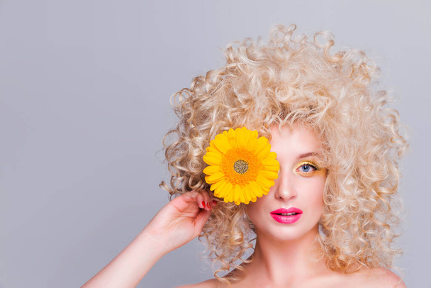 Красива модна блондинка з об'ємною кучерявою зачіскою, голими плечима і соняшником в руках на сірому фоні. жінка дивиться на камеру і прикриває око квіткою
 - Фото, зображення